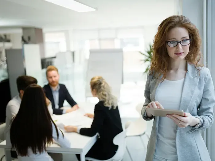 Professional woman with tablet near glass meeting room as colleagues discuss strategy at table in a modern office